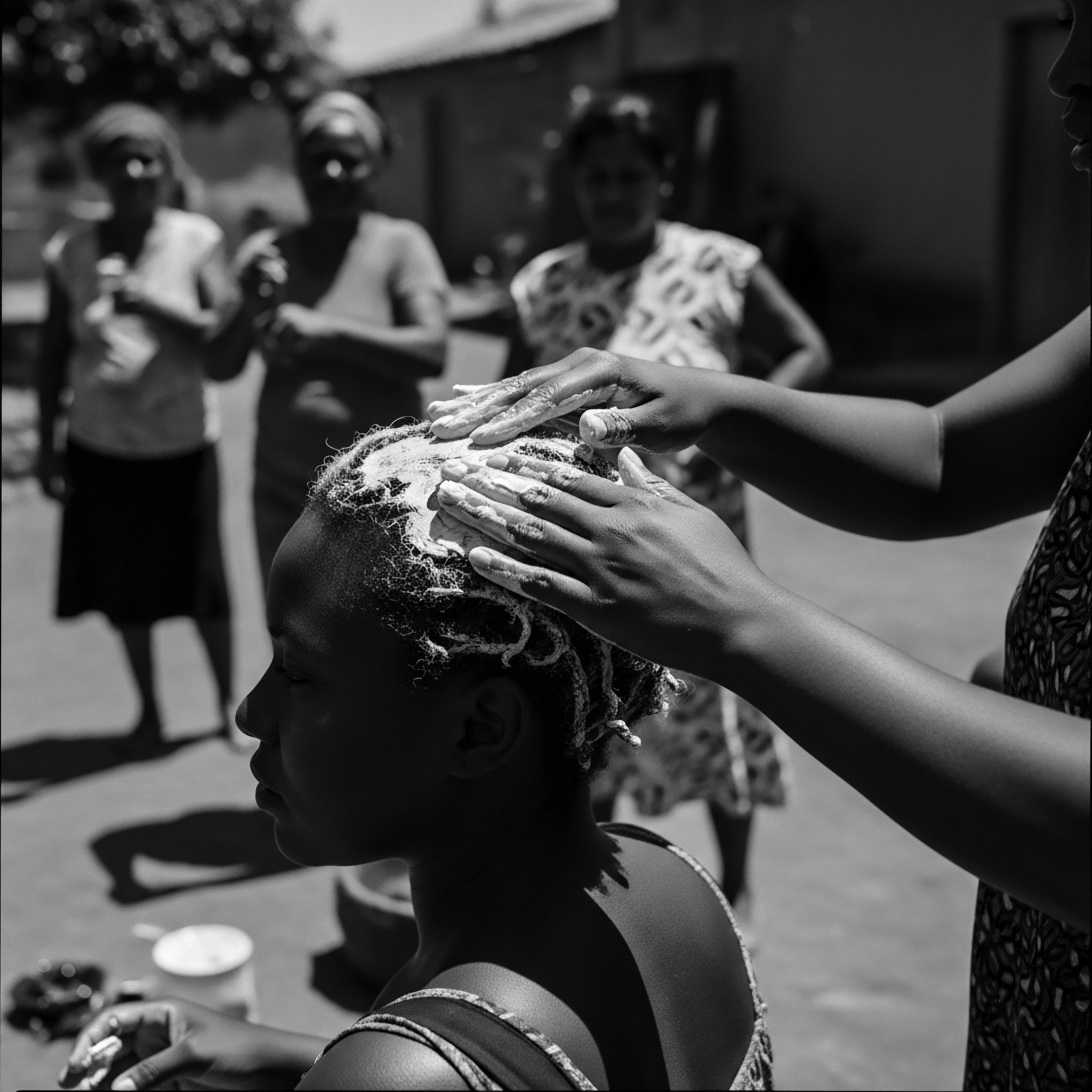 Black and White Image of Textured Hair Treatment Ritual Outdoor Setting Hands administer creamy treatment to textured coils, as women stand by, witnessing an outdoor hair ritual rooted in ancestral heritage and holistic wellness practices for Black hair the scene offers a poignant reflection on historical hair care traditions passed down through generations, emphasizing the importance of heritage and community.