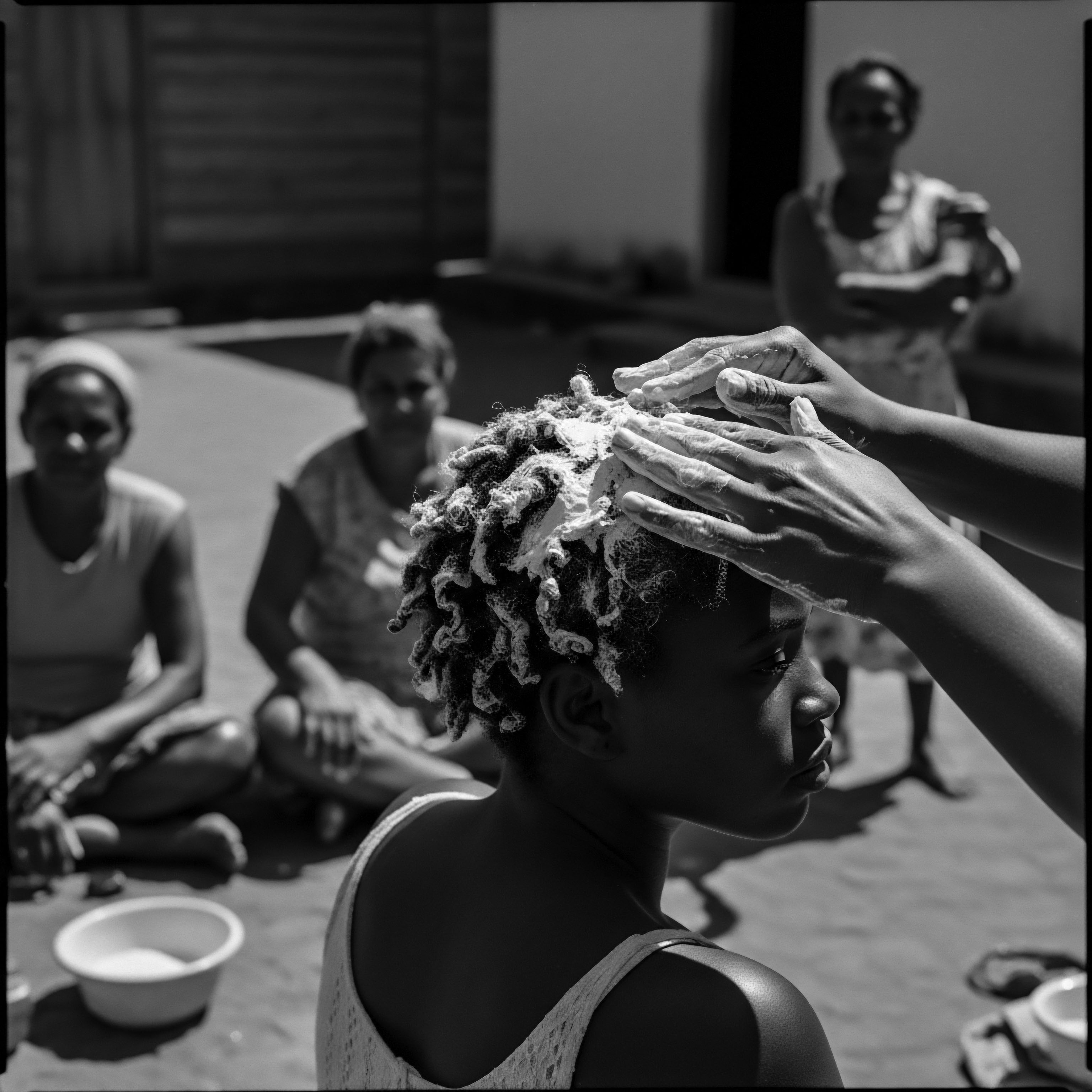 Black and White Image of Young Woman Receiving Scalp Treatment Outdoors Hands gently massage a scalp treatment into tightly coiled hair, amidst onlookers, symbolizing a deep connection to heritage and holistic self-care. The black and white aesthetic underscores the timelessness of these ancestral practices, reflecting the enduring beauty standards and communal bonds associated with textured hair.