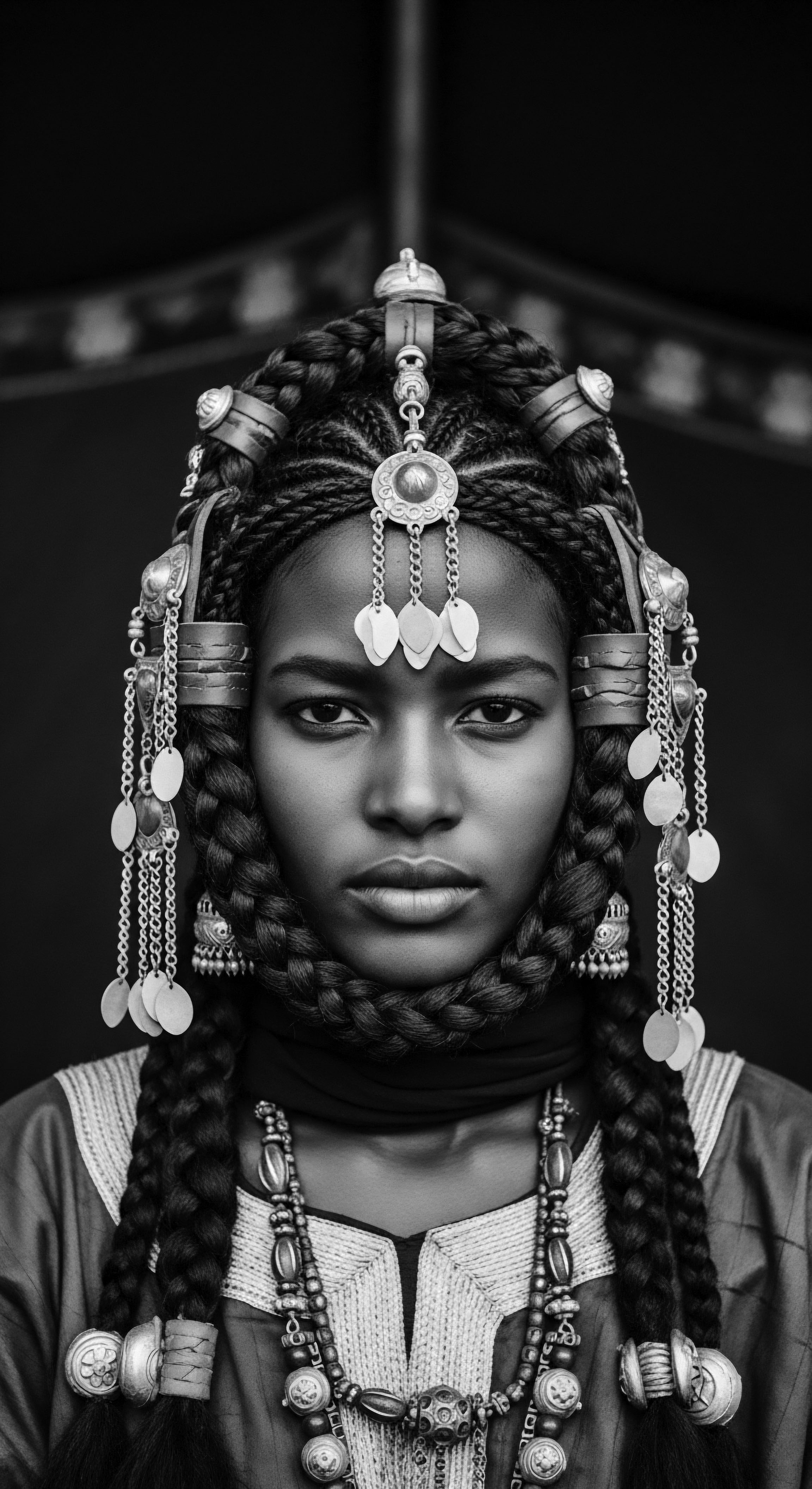Black and White Portrait of Woman Adorned in Braids and Jewelry Intricate braiding designs and silver adornments showcase a rich cultural heritage within this woman's portrait. Her striking gaze and composed expression, complemented by the meticulous detail of her textured hair and jewelry, invites contemplation on identity, ancestral bonds, and expressive styling.