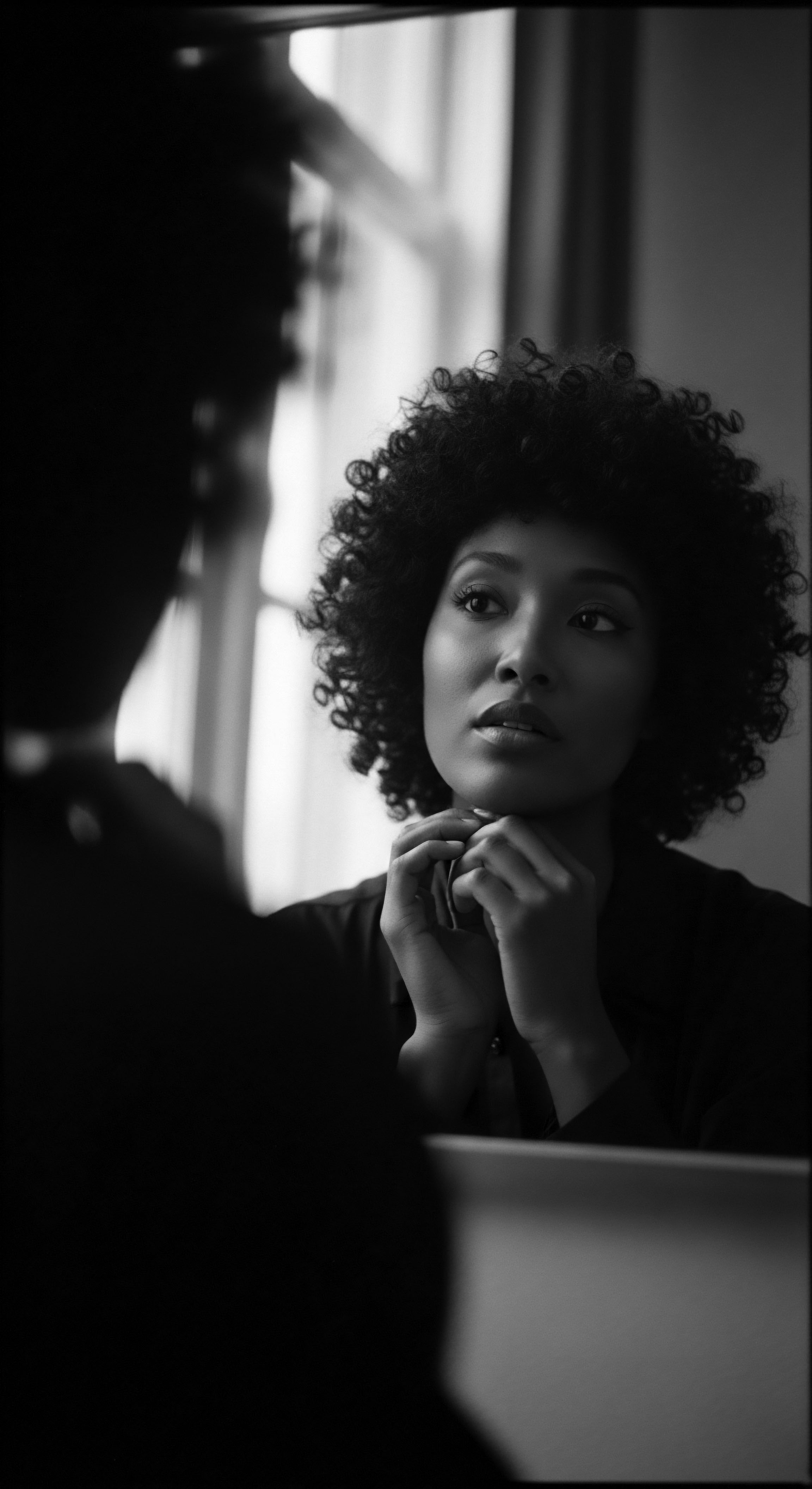 A monochrome image reveals a woman with a voluminous afro, her hands gently supporting her chin. The soft light accentuates her textured hair's coil patterns and subtle skin tones. Reflected in a mirror, the visual composition speaks to self-perception and the timeless beauty found in natural black hair traditions.