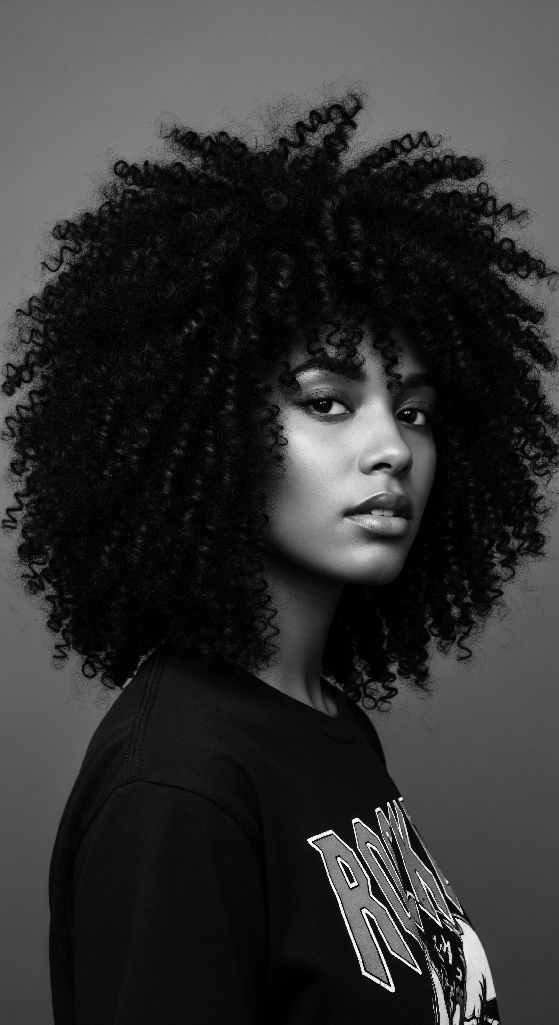 Elegant Black Woman with Natural Afro Coils in Dramatic Monochrome Portrait This evocative monochrome portrait captures the essence of afro coiled beauty, reflecting a legacy of ancestral heritage. The rich textures and the subject's striking gaze invite contemplation on identity and self-expression through natural coiled hair, a powerful symbol of cultural pride and conscious holistic care.