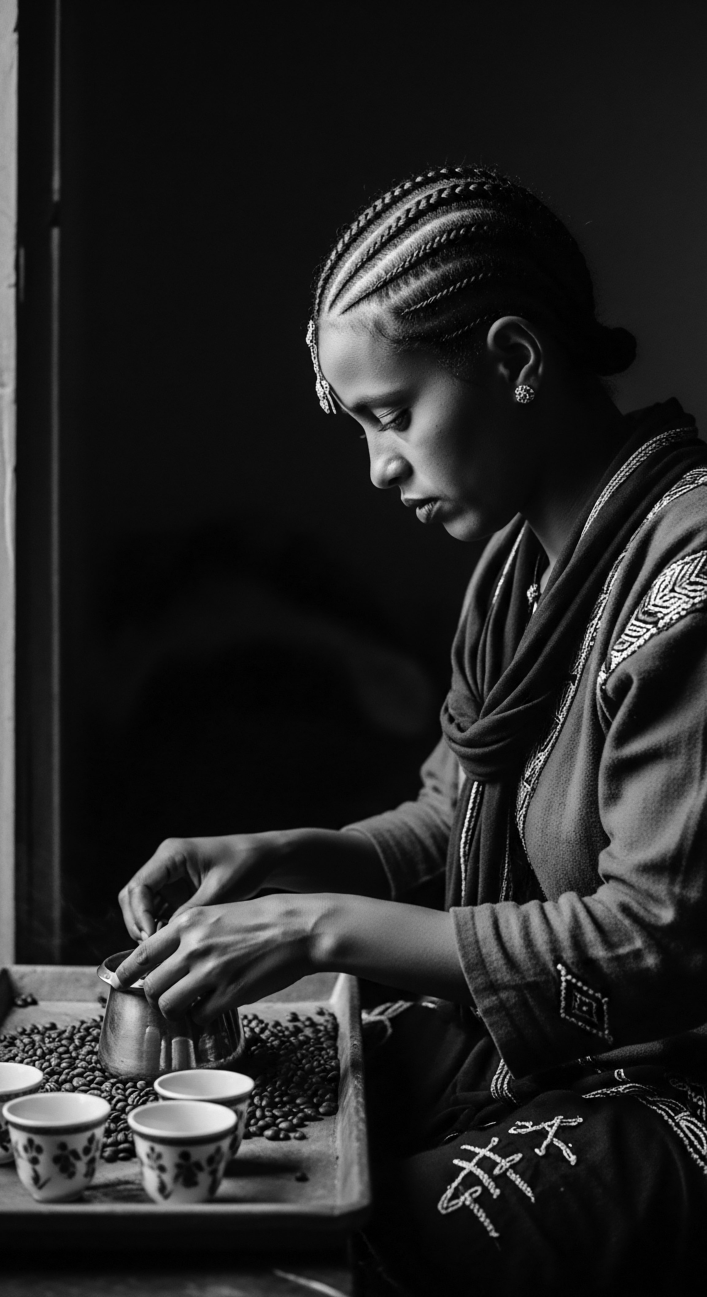 Captured in stark monochrome, the image tells of Ethiopian heritage through a woman preparing coffee with tightly coiled braids, adorned in traditional jewelry, a cultural ritual reflecting deep roots and beauty with expertly crafted detail, celebrating ancestral traditions and holistic hair care.