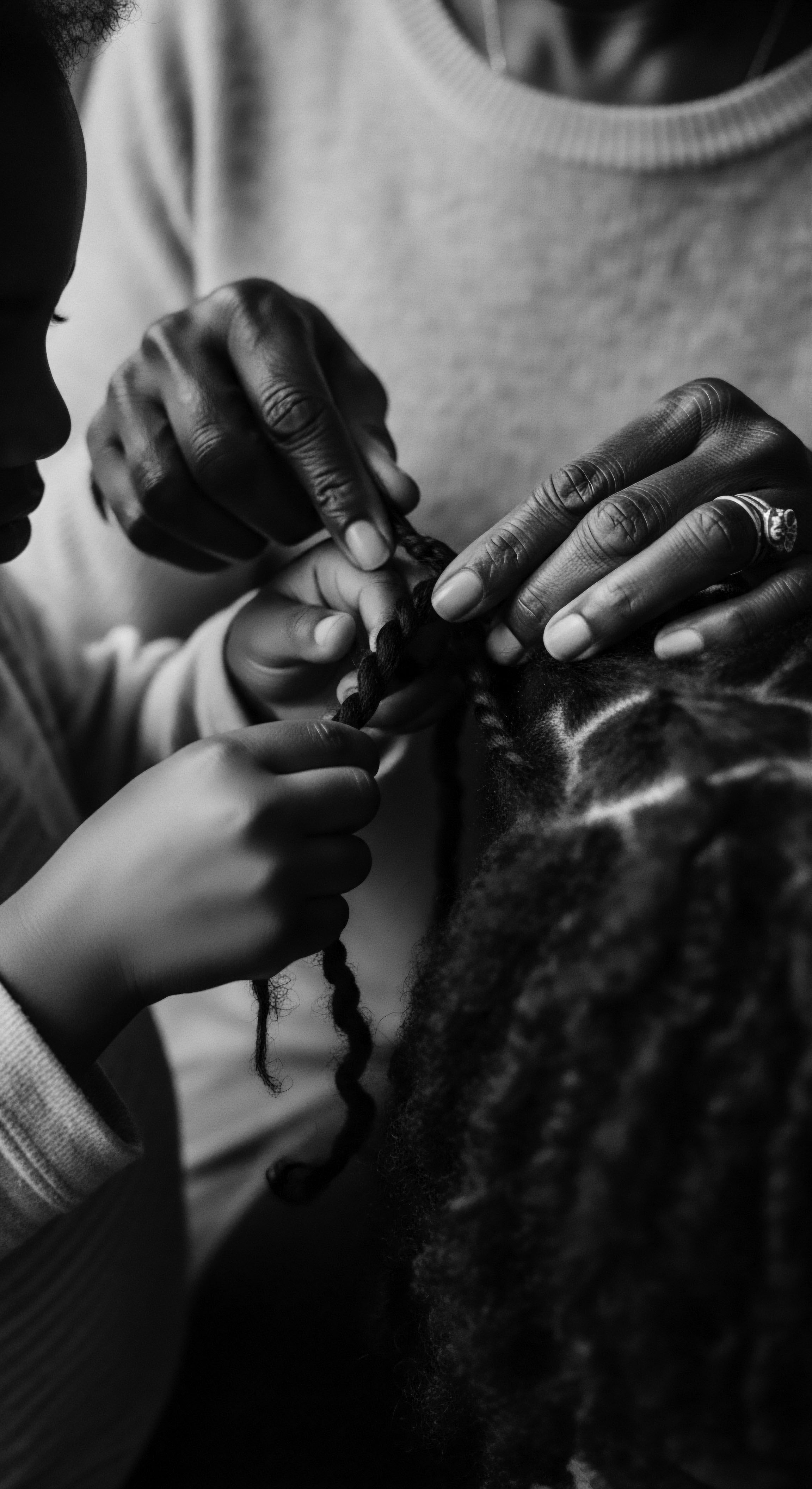 Generational Hands Braiding Black Textured Hair: Ancestral Tradition and Natural Care A black and white image resonates deeply through showcasing the passing down of cultural knowledge via hands intertwining kinky hair. This familial moment celebrates heritage, highlights the intricate artistry of black hairstyling traditions, and emphasizes commitment to natural hair care within an intergenerational black family dynamic, enhancing porosity.