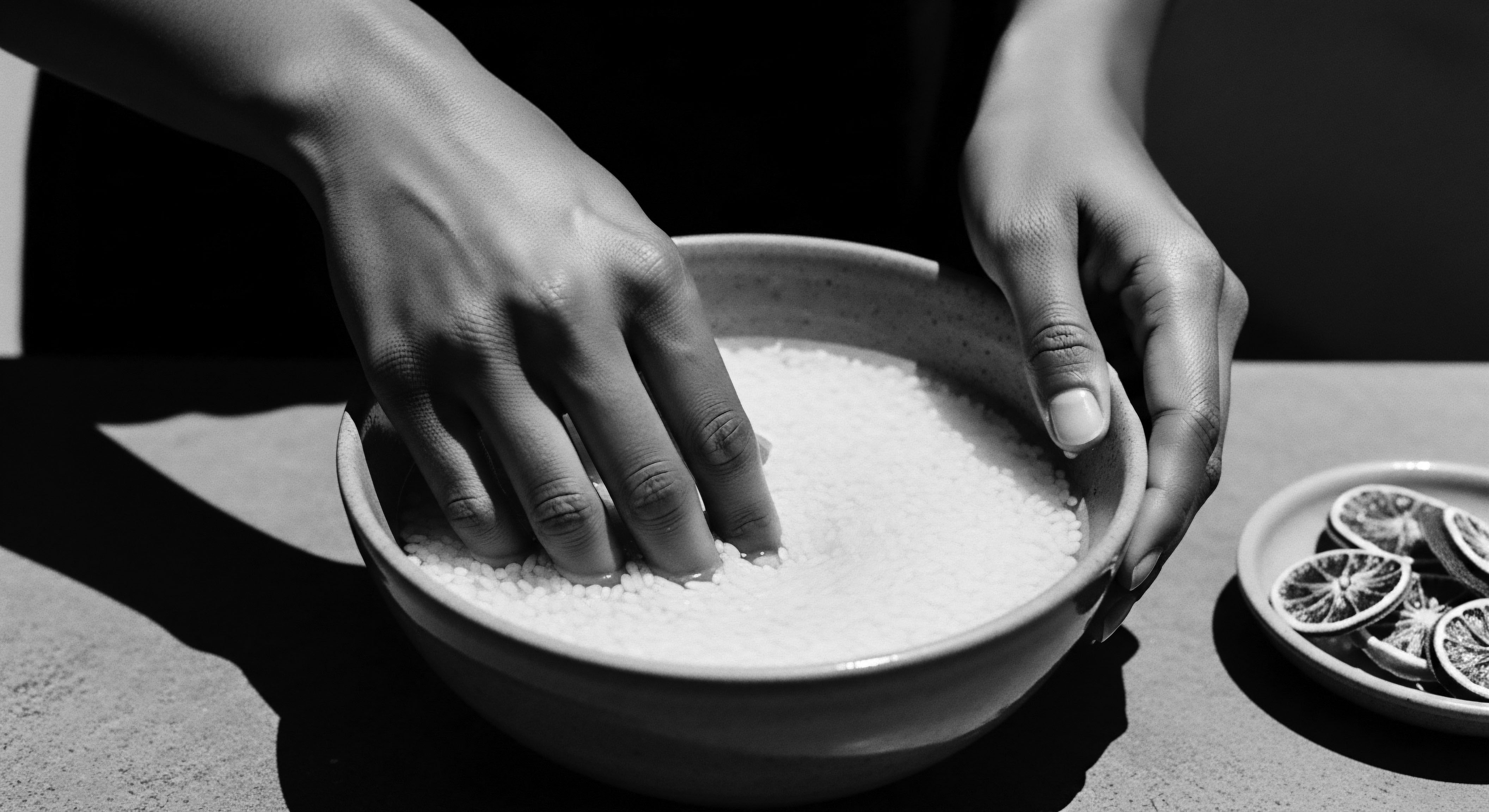 Hands Gently Immersing Rice Grains Water Bowl Black and White Still Life Hands immersed in rice water embody a connection to generations past, celebrating its traditional use in clarifying and softening skin. This holistic practice honors ancient rituals, enhancing the beauty of melanated skin and highlighting the significance of natural elements in ancestral care.