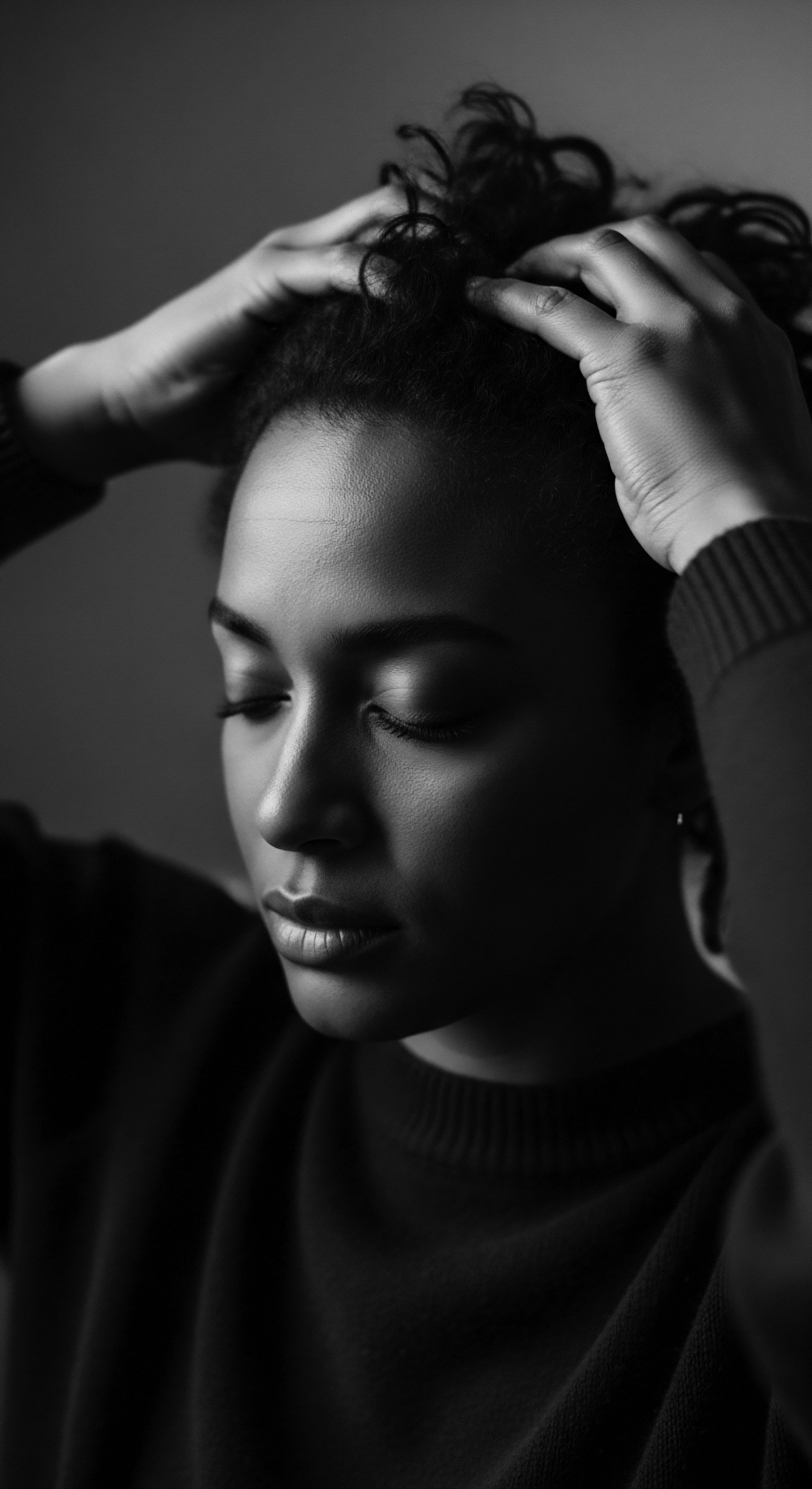 Introspective Portrait Woman with Coiled Hair Hands Framing Face The black and white image captures a moment of quiet contemplation, as the woman's hands rest upon her textured coiled hair formation. The intimate scene suggests a connection to heritage, hair wellness traditions, and personal identity interwoven through care and styling techniques rooted in ancestral and holistic methodologies.