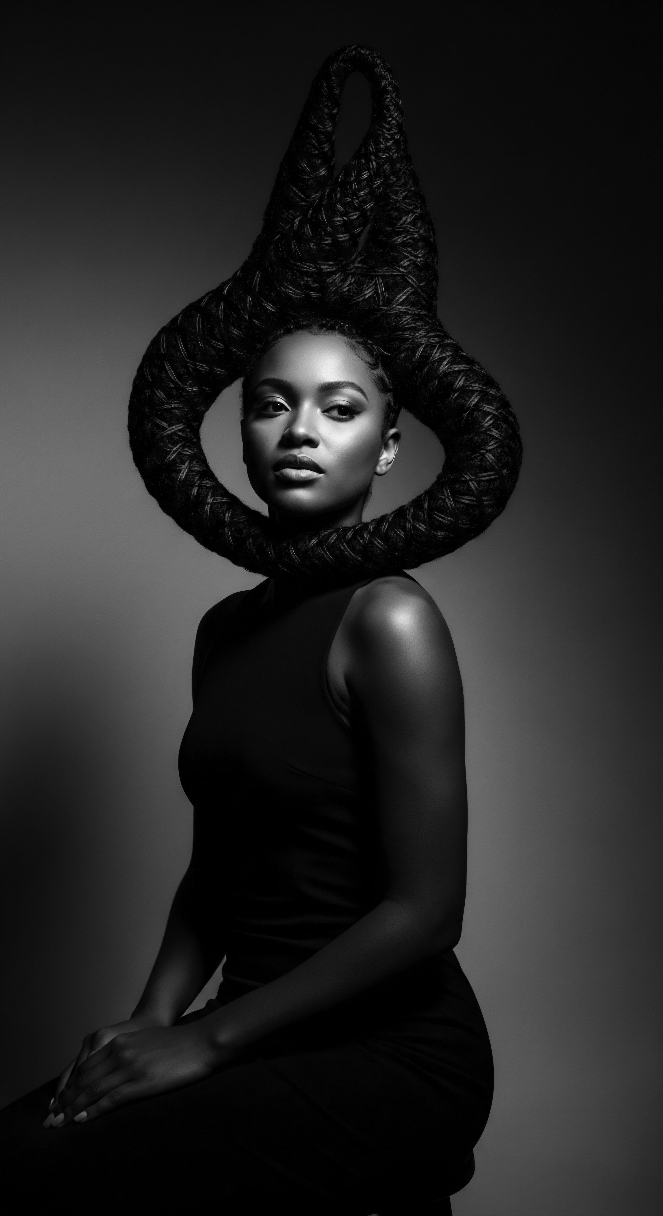 A striking black and white portrait presents a woman of color wearing a sleek black dress, her hands gently clasped. Dramatic lighting accentuates her complexion and the elaborate braided hairstyle, a circular halo emphasizing hair texture and a creative expression of identity and ancestral aesthetics.
