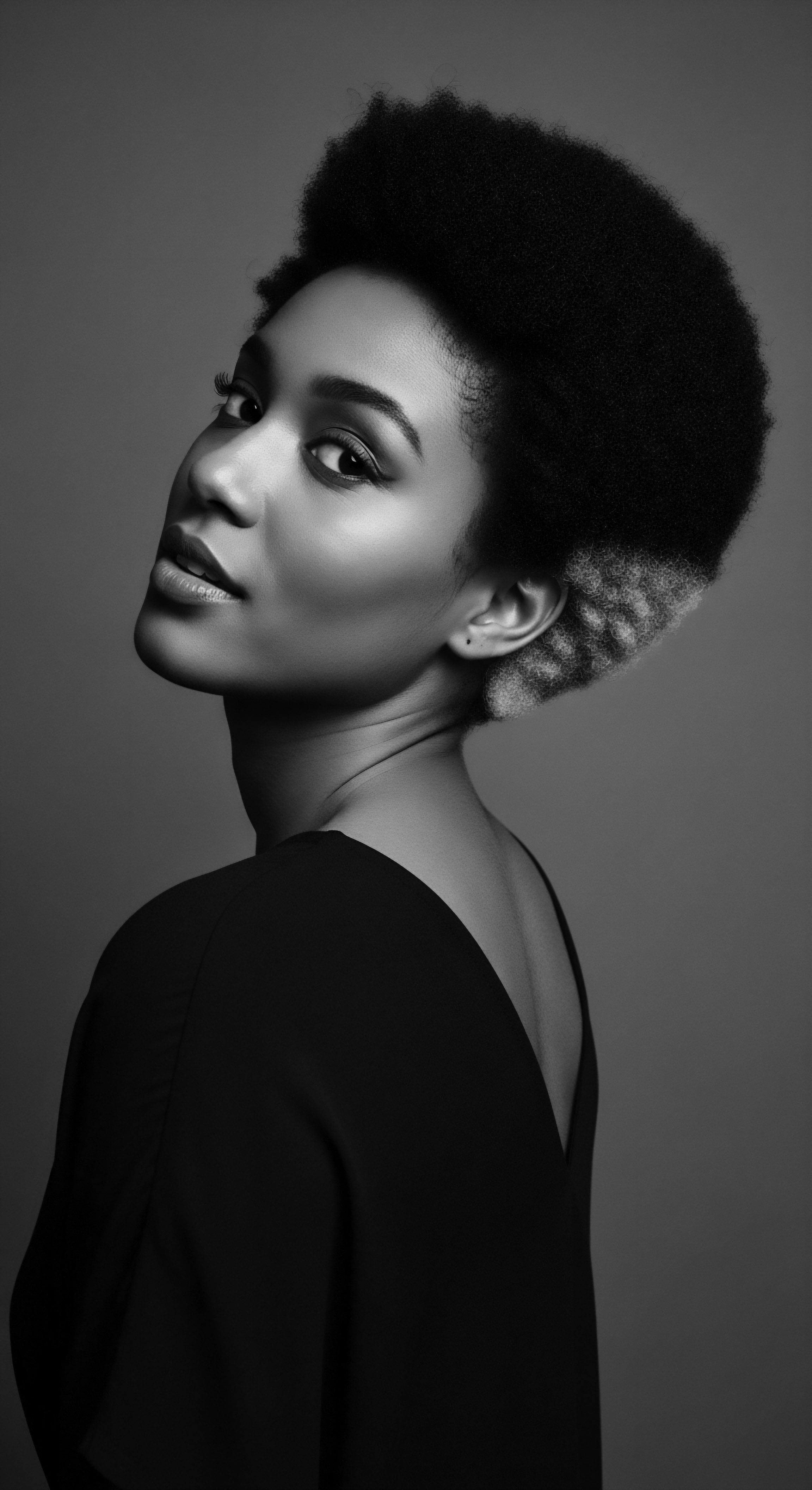 Elegant black and white portrait showcases a Black woman with a striking afro, featuring a short tapered silhouette. The textured hair presents a bicolor contrast, accentuating natural volume. Dramatic lighting highlights facial contours, emphasizing the beauty and texture in heritage inspired expressive styling.
