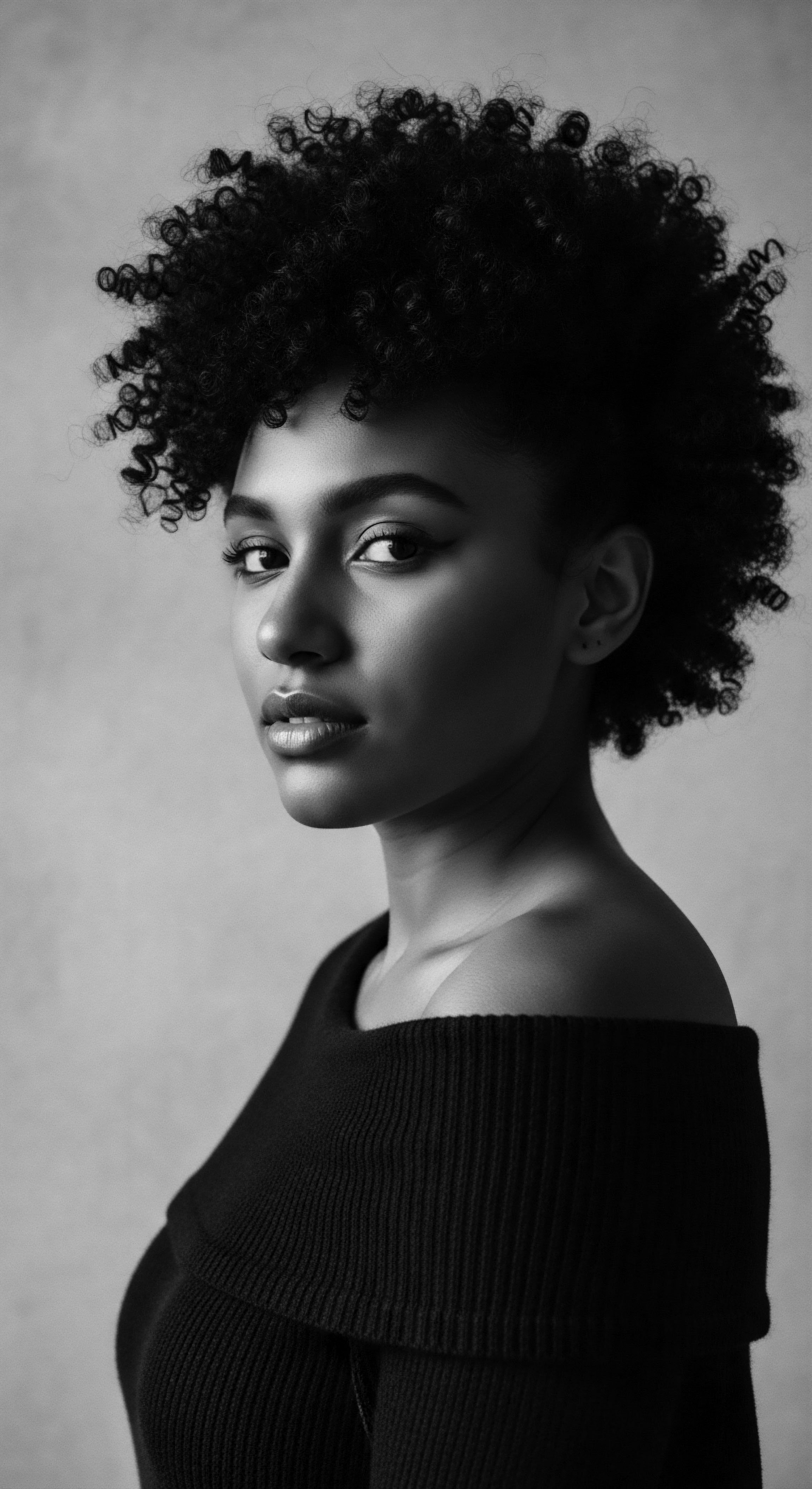 Monochrome Portrait of a Woman Displaying Afro Textured Hair Heritage Monochrome artistry highlights the beauty of afro textured hair, styled with volume and bold expression. The confident gaze, paired with the off-shoulder top, honors the heritage and expressive potential of natural afro textured hair. This image is an ode to self assured beauty.