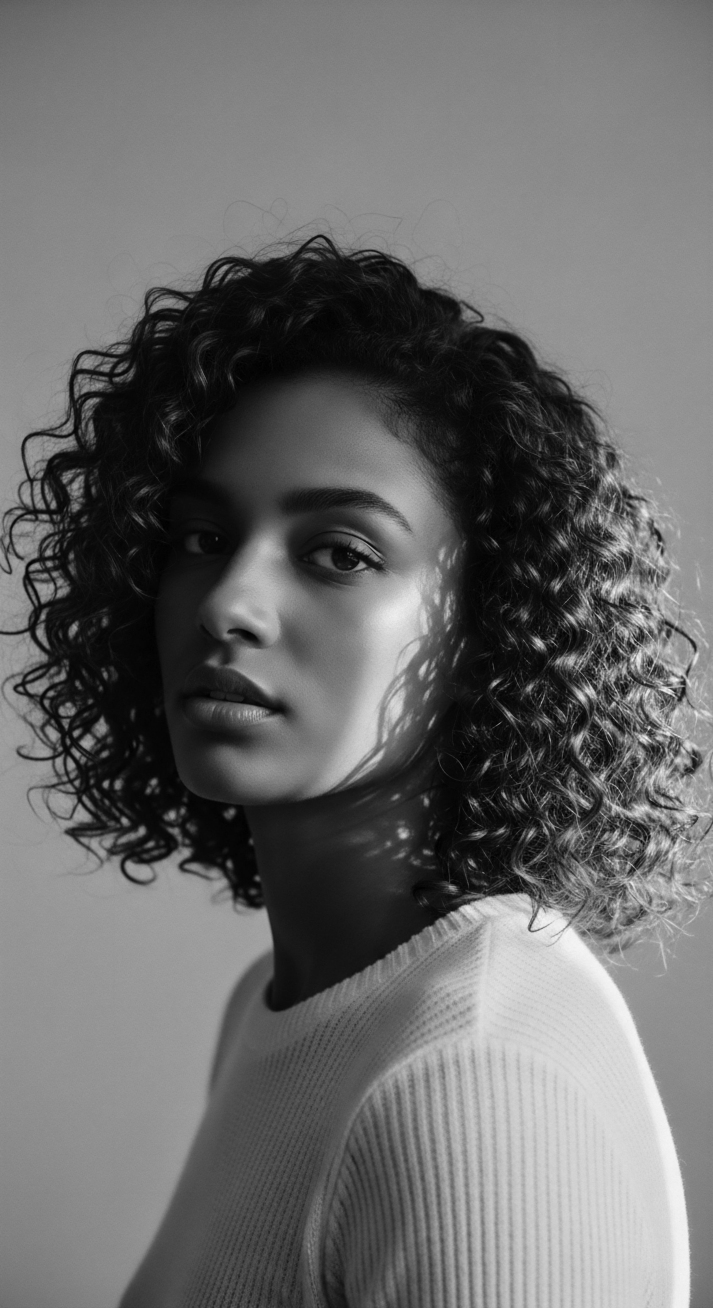 The monochrome tones accentuate the beauty and resilience in her tightly coiled hair, a textured crown that reflects a powerful legacy. Light and shadow sculpt a portrait that invites contemplation on ancestral roots, identity expression, and the art of holistic hair care traditions.