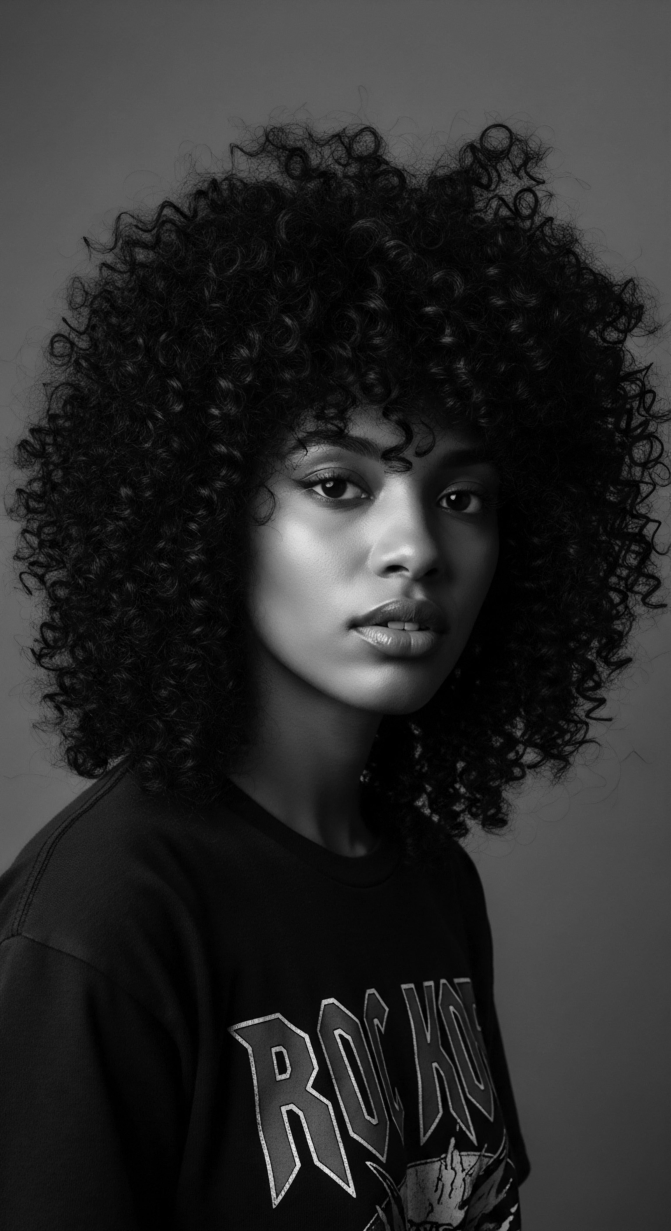 The monochromatic study highlights the intricate texture of Afro hair as a form of cultural expression. The woman's confident posture and gaze amplify this message, inviting viewers to contemplate the enduring significance of naturally coiled hair in identity narratives and ancestral heritage.