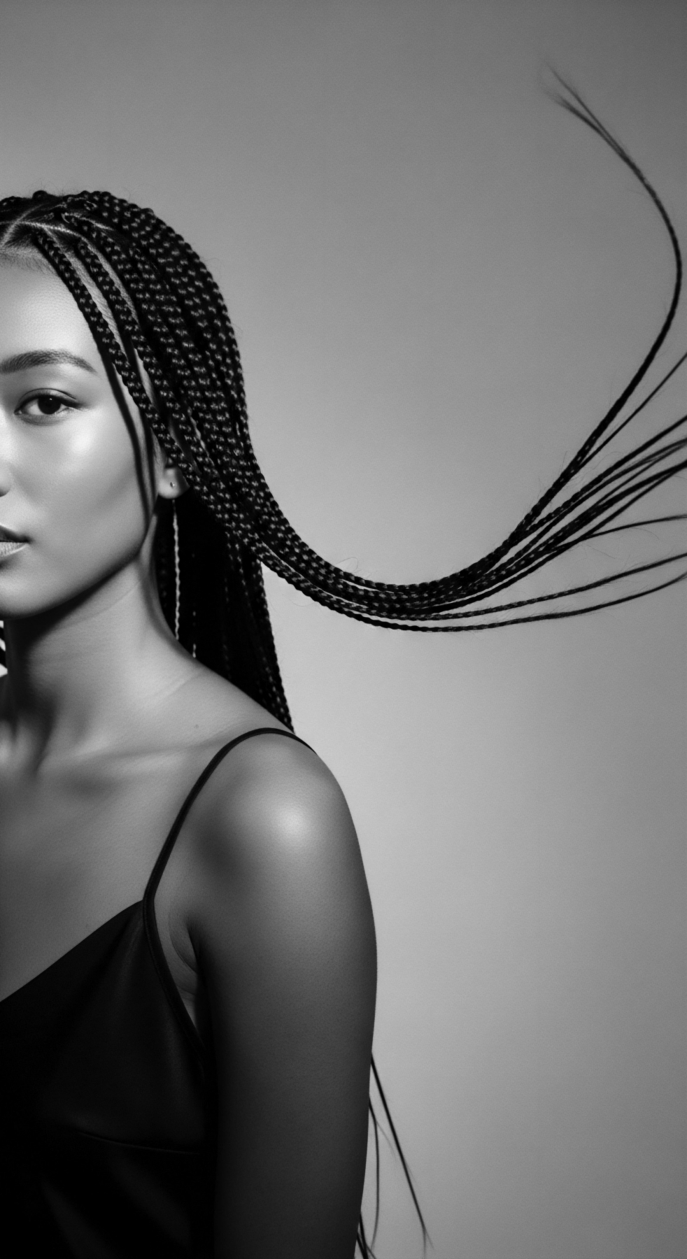 This black and white portrait showcases a woman's beauty, highlighting her braids that cascade dynamically against a neutral backdrop. Soft light emphasizes skin texture and intricate braid detailing. The composition captures heritage and modern style, celebrating unique textured hair.