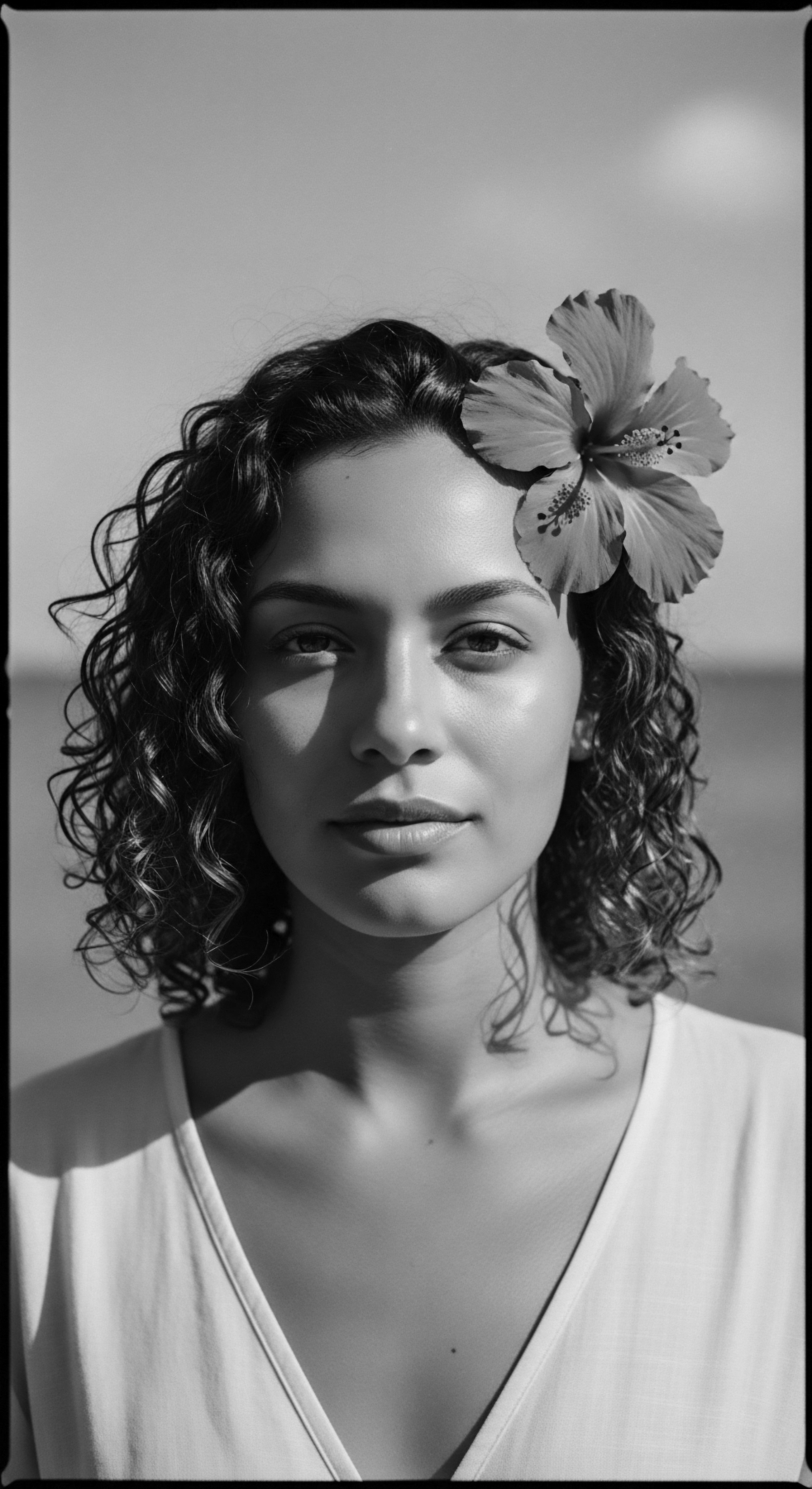 A captivating monochrome portrait features a woman with shoulder-length textured hair demonstrating soft undulation and a radiant hibiscus bloom accenting her crown. The interplay of natural light and shadow enhances her features, invoking timeless beauty and ancestral heritage with expressive styling, while reflecting wellness.