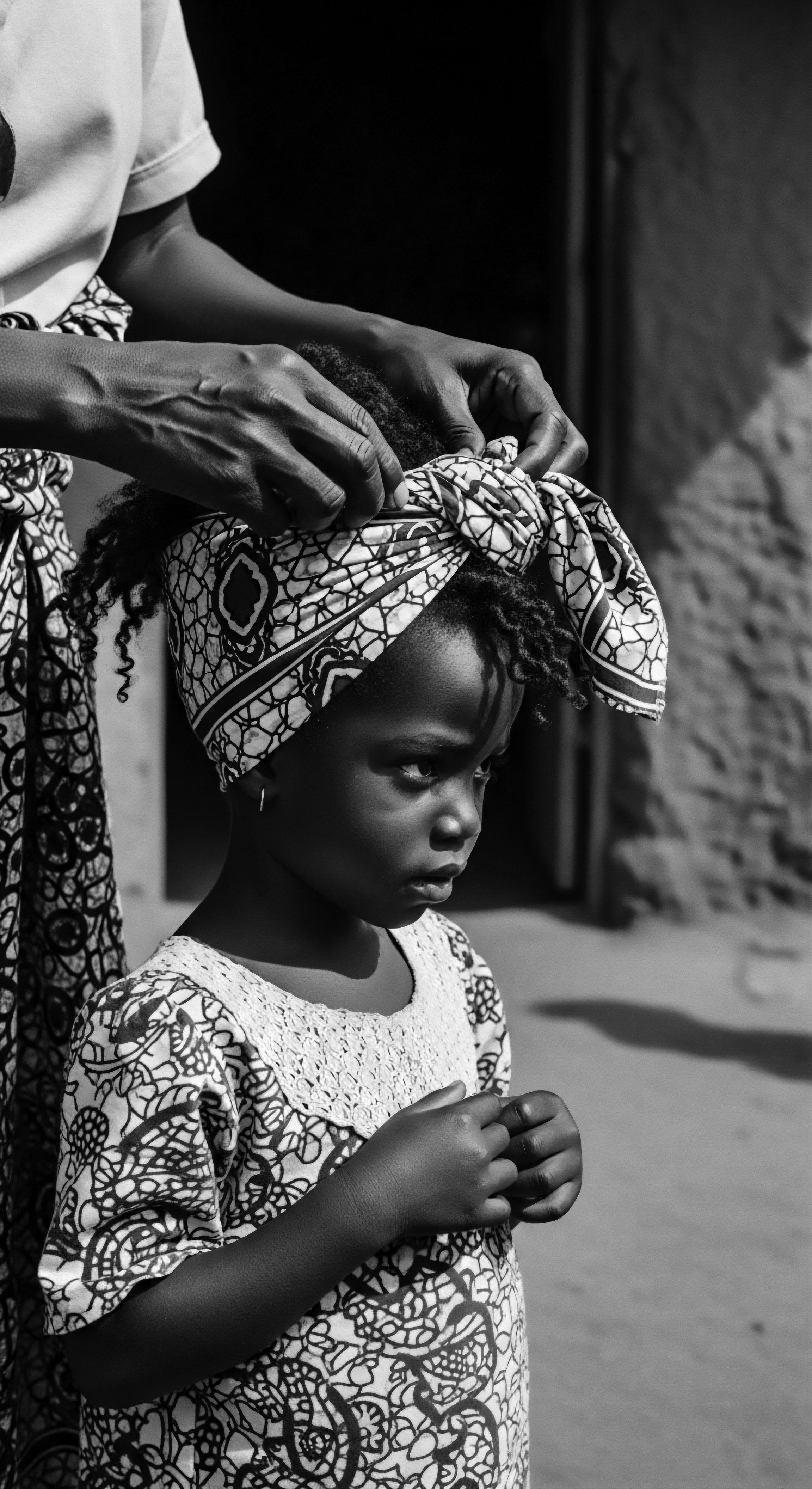 Mother Tying Head Scarf on Young Child Black and White Portrait The striking monochrome aesthetic underscores a generational bond as a mother carefully secures a traditional headscarf on her child's textured coils. This intimate act visually celebrates cultural identity, ancestral heritage, and the enduring artistry expressed through Black hair traditions and expressive styling.