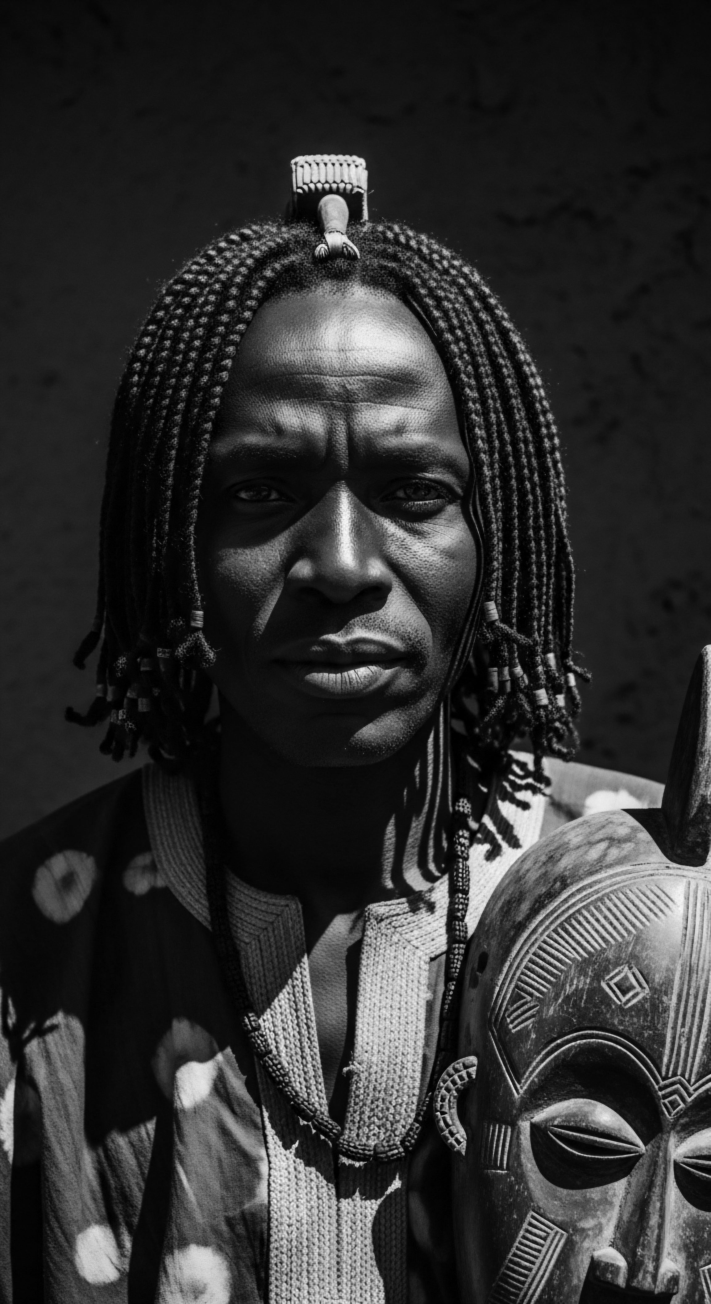 A close-up black and white portrait reveals a man with intricately braided hair adorned with beads, standing against a stark backdrop. He holds a carved wooden mask reflecting ancestral heritage. The image explores themes of cultural identity, captured through detailed textures and the interplay of light and shadow on both the skin and the braiding patterns.