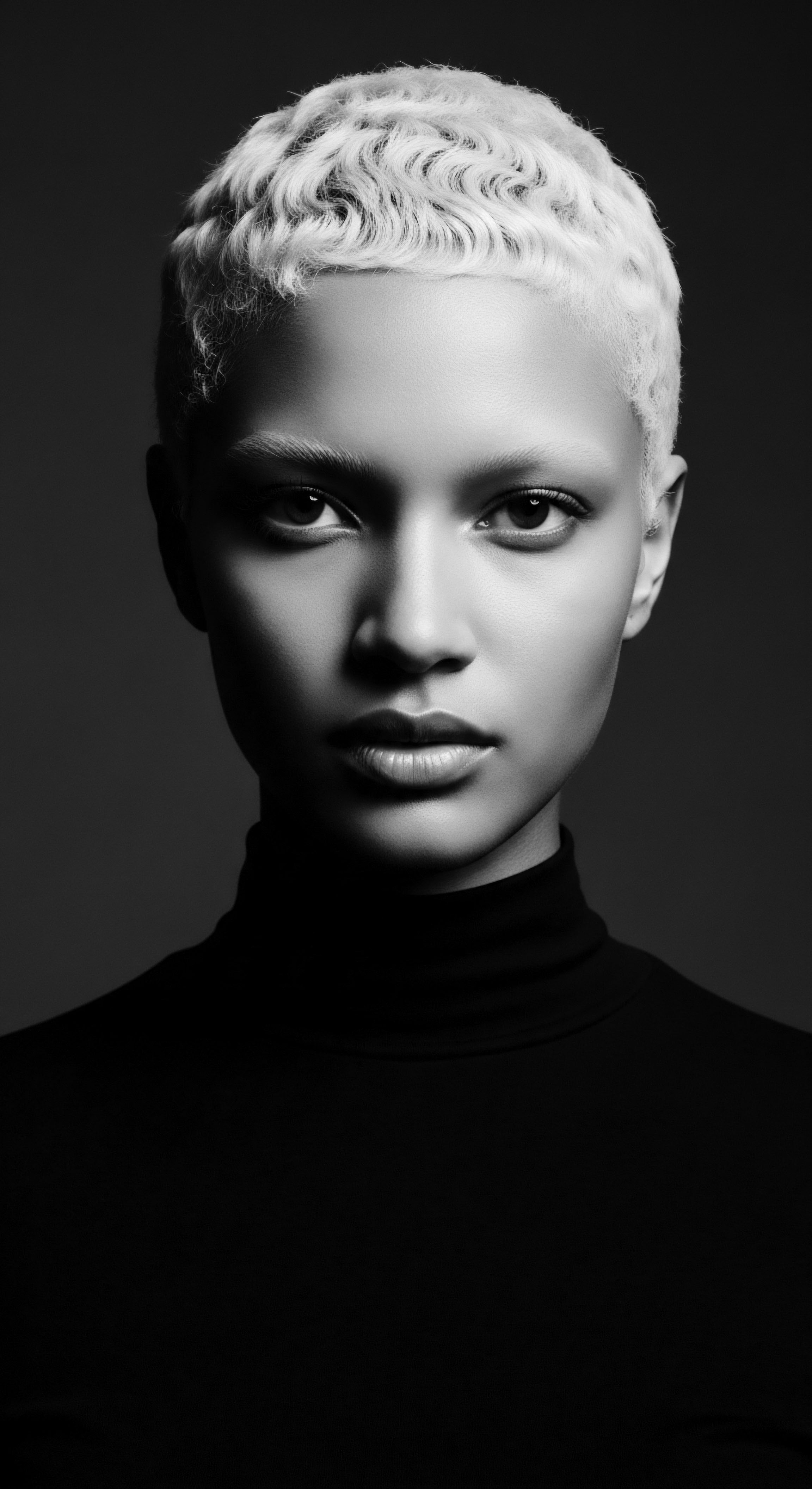 The interplay of light and shadow accentuates the platinum blonde, short, textured hair's unique wave pattern, framing her direct gaze in a study of minimalist portraiture. This visual exploration uses monochrome to emphasize heritage, striking features and an intimate sense of self-expression through textured hair.