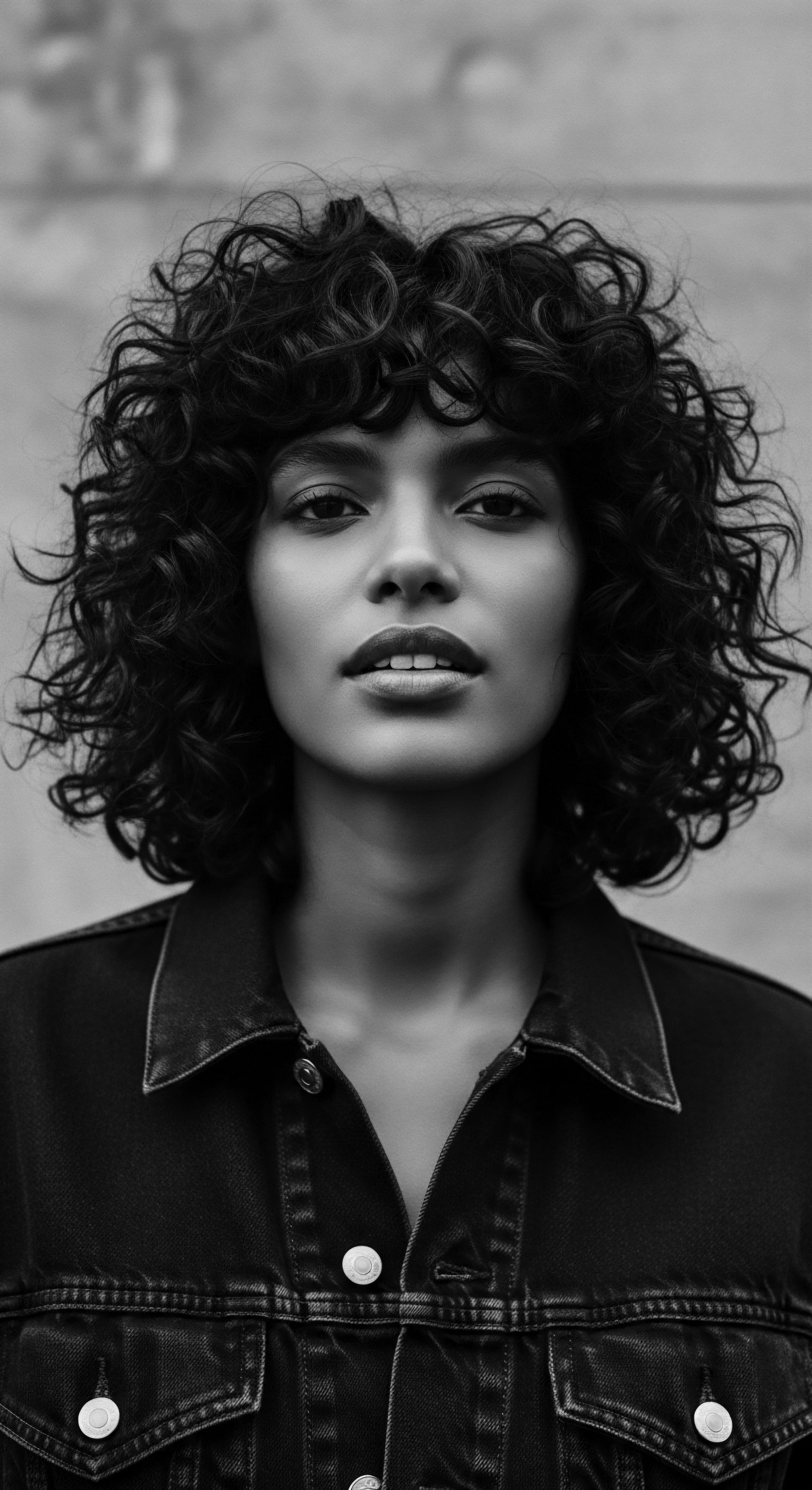 Striking Portrait of a Woman with Bold Coiled Afro and Denim Jacket This striking black and white portrait showcases the power of authentic self-expression through natural coiled hair, styled boldly to accentuate her features and heritage, while the denim jacket brings a contemporary edge, uniting ancestral pride with a modern aesthetic.