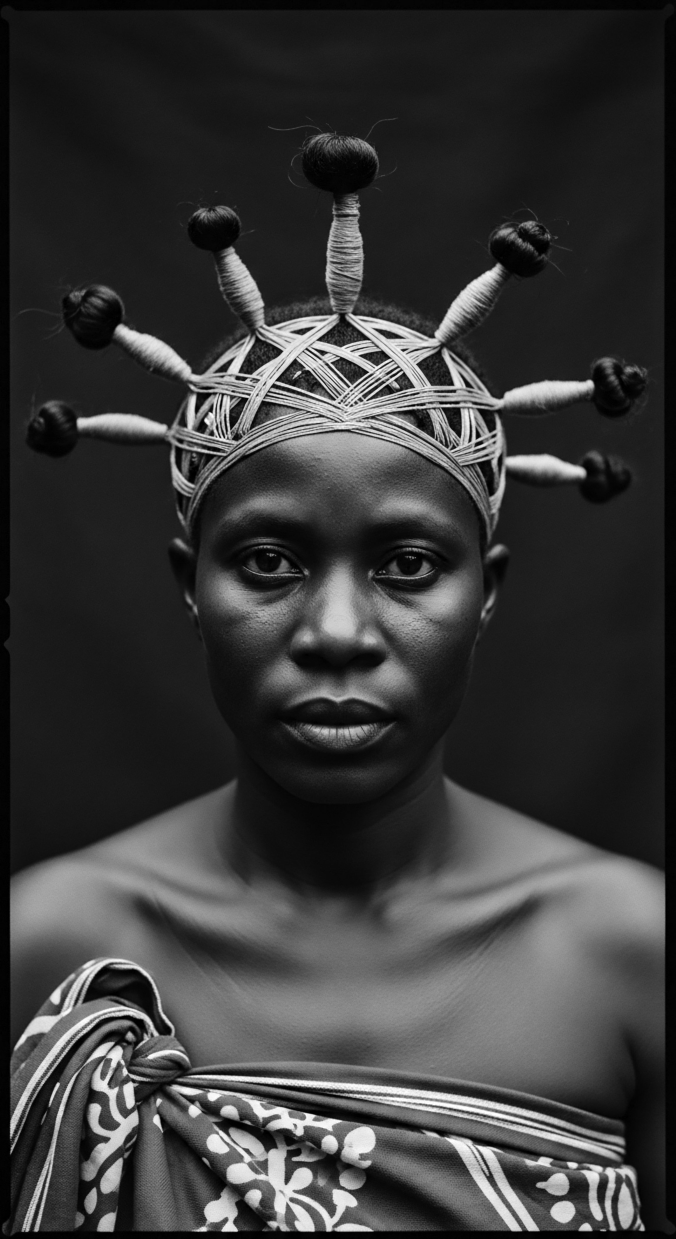 Striking Portrait of Woman Adorned with Intricate Braided Hairstyle and Headpiece The monochrome portrait highlights textured hair's artistry in an elaborate braided updo, where wrapped extensions radiate like a crown. A patterned cloth adds a layer of ancestral heritage, while light and shadow delicately define the woman's facial features, inviting contemplation of identity and cultural pride through expressive styling.