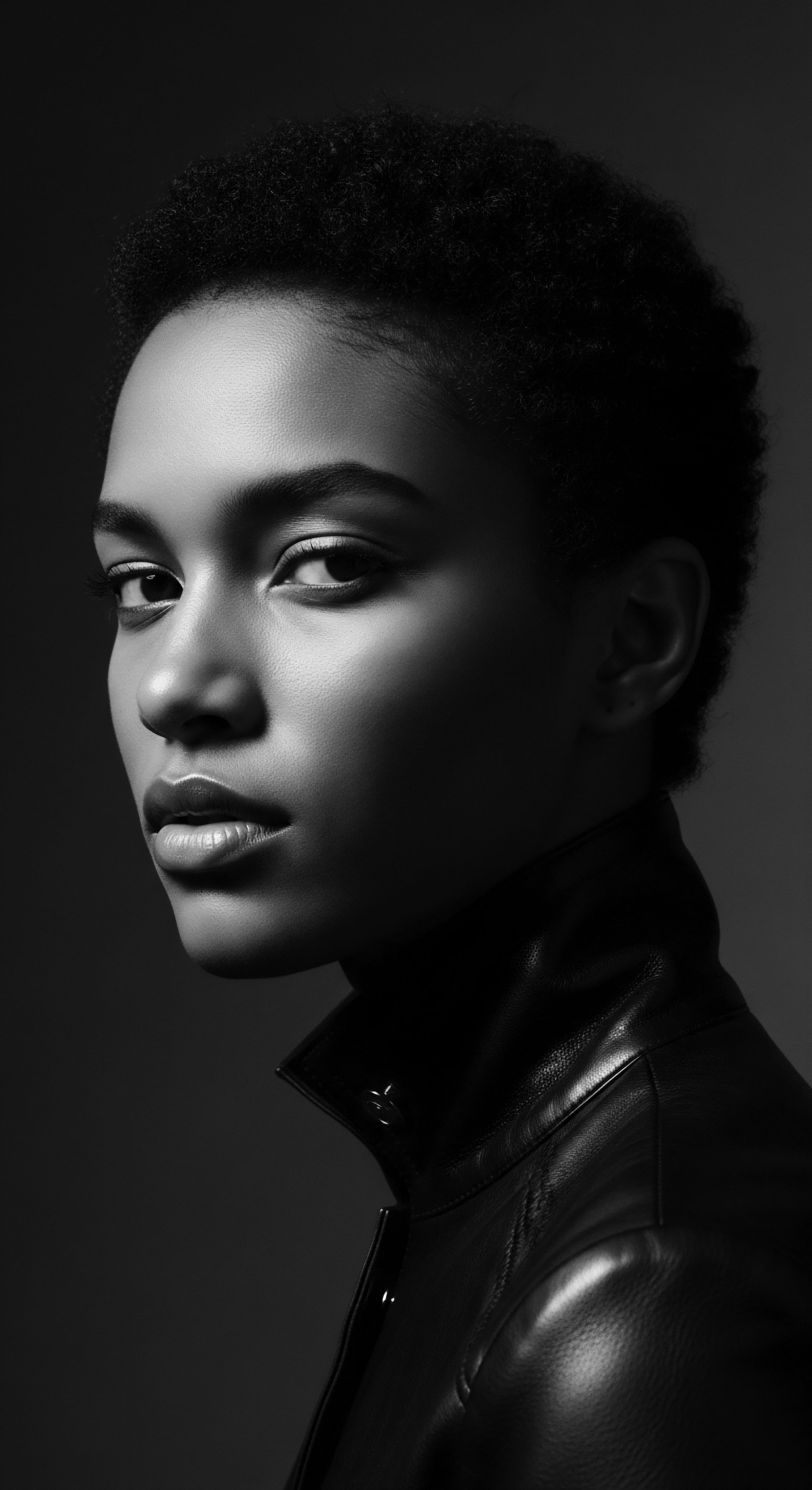 Striking Portrait of Woman with Afro Textured Hair in Leather Jacket This image celebrates the elegance and power of afro hair, highlighting its natural texture and the confident beauty of its wearer. The contrast of light and dark enhances the inherent sophistication of her style, reflecting ancestral heritage and modern expression.