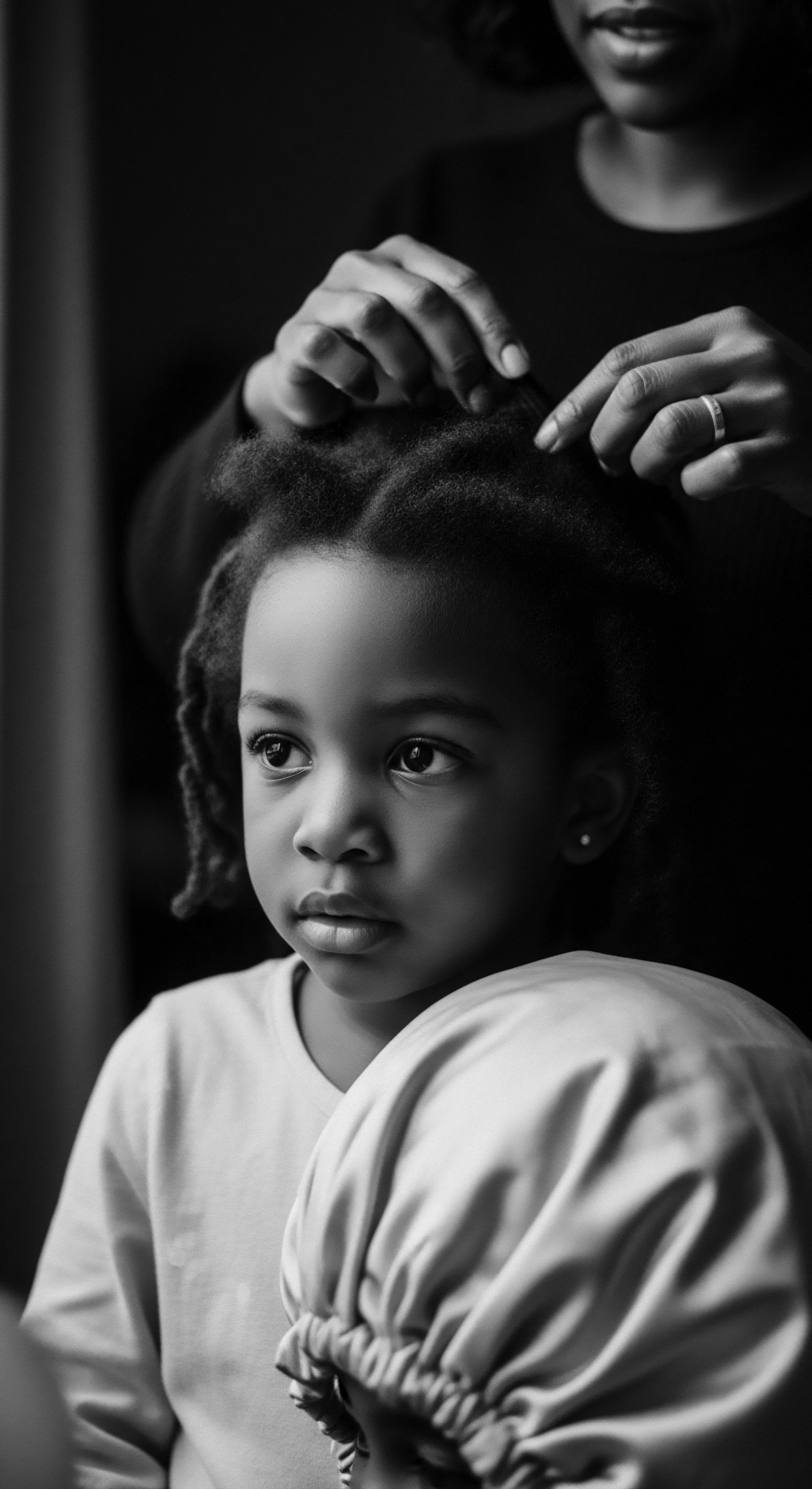 A serene black and white portrait captures a mother gently styling her daughter's textured hair, forming intricate braids. Soft light illuminates the child's thoughtful expression, set against a dark backdrop. The image celebrates cultural heritage, intimate bonding, and the care involved in managing natural hair formations.
