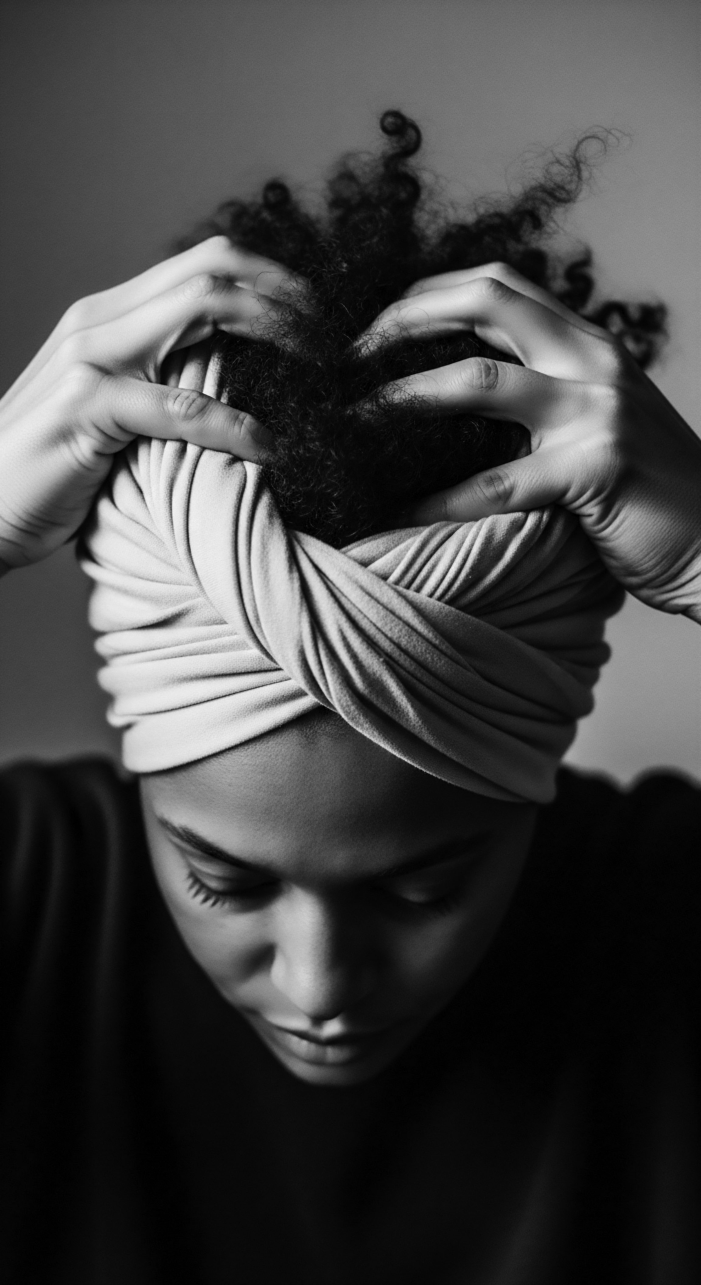 Woman Embracing Natural Hair Texture with Protective Headwrap in Monochrome The photograph honors the intimate ritual of textured hair care, as seen in the artful arrangement of the headwrap and the gentle touch, symbolizing connection to heritage, self-expression, and the embrace of natural beauty through protective styling practices and mindful, holistic self-care traditions.