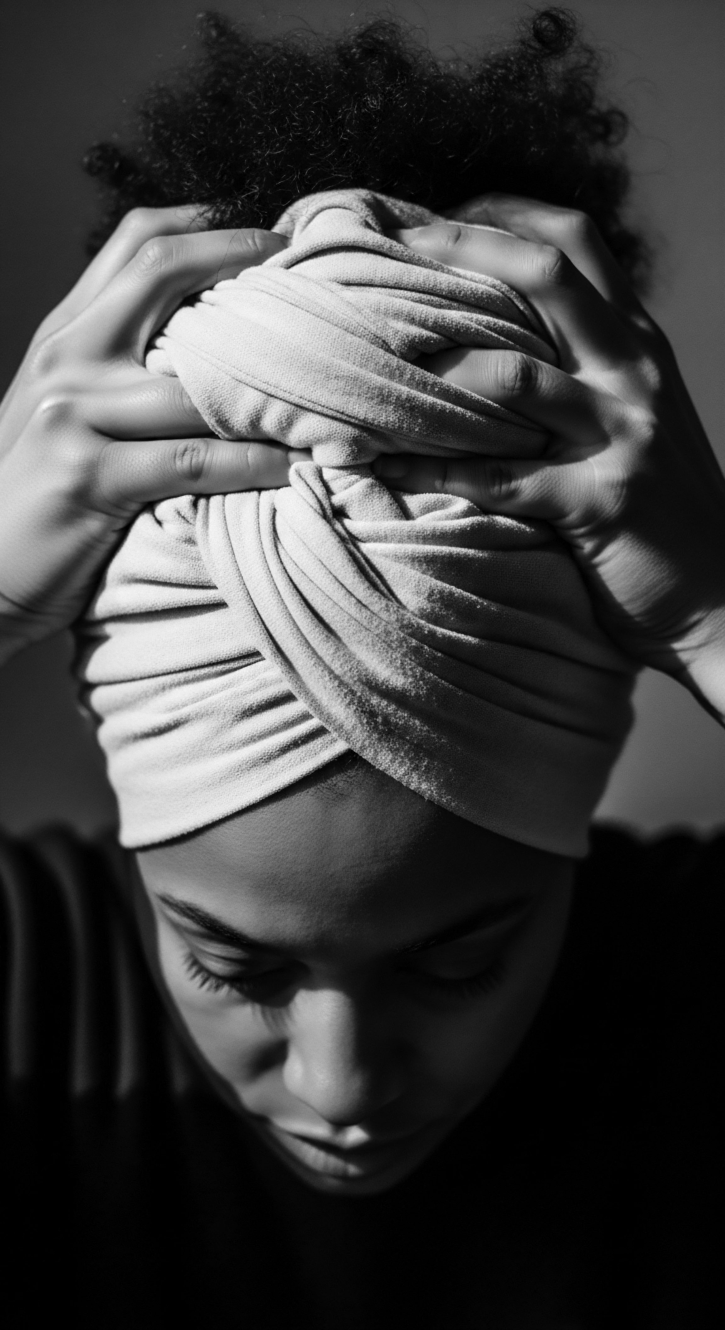 The textured hair traditions are beautifully embraced as a woman carefully arranges a turban, the interplay of light and shadow signifying a moment of holistic wellness, deep connection to ancestral roots, and expressive self care, emphasizing the elegance and heritage within Afro hair practices.