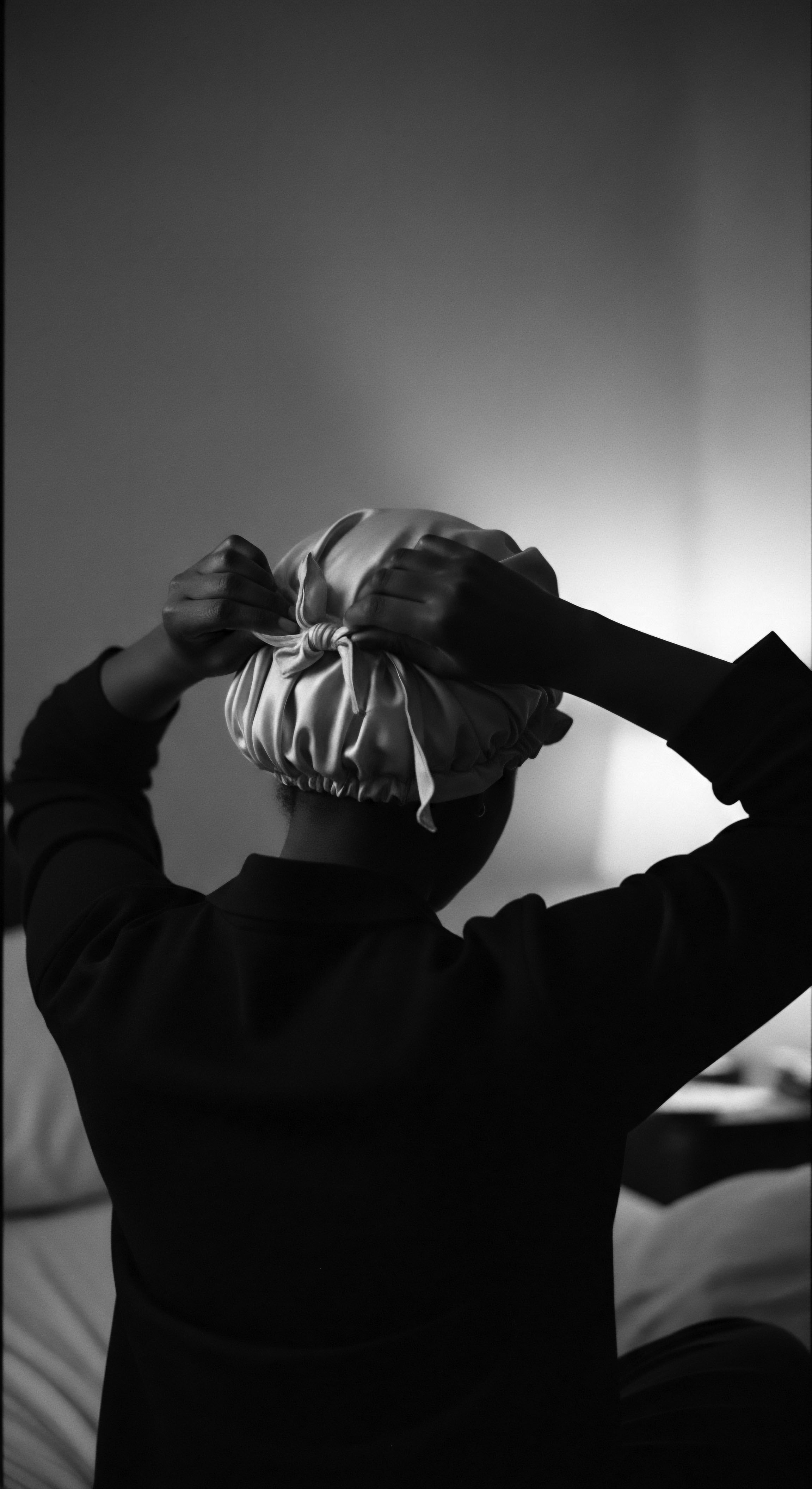 Woman Securing Silk Bonnet Overnight Hair Protection Black and White Photo The monochrome image captures a nightly self-care ritual, securing a silk bonnet to protect textured hair and preserve moisture, reflecting ancestral heritage and Black hair traditions rooted in maintaining healthy, well-hydrated coils, promoting wellness and embracing the natural beauty of coiled formations.