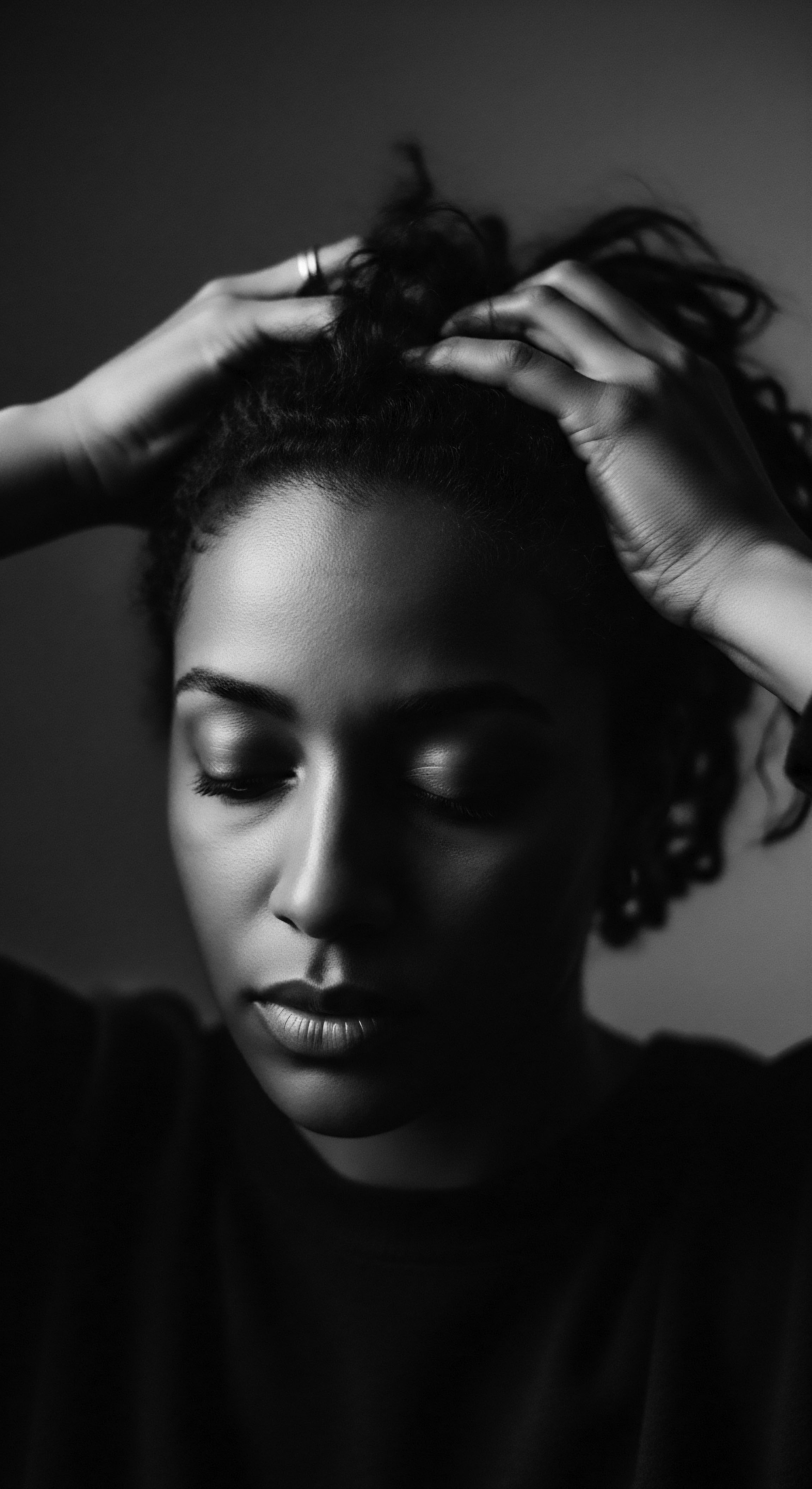 Woman with Closed Eyes Touching Coiled Hair in Black and White Hands gently caressing textured coils, she embodies self-reflection, a quiet moment honoring ancestral heritage and nurturing holistic well-being. The interplay of light and shadow highlights the beauty of natural hair, emphasizing a spiritual connection through mindful care practices.