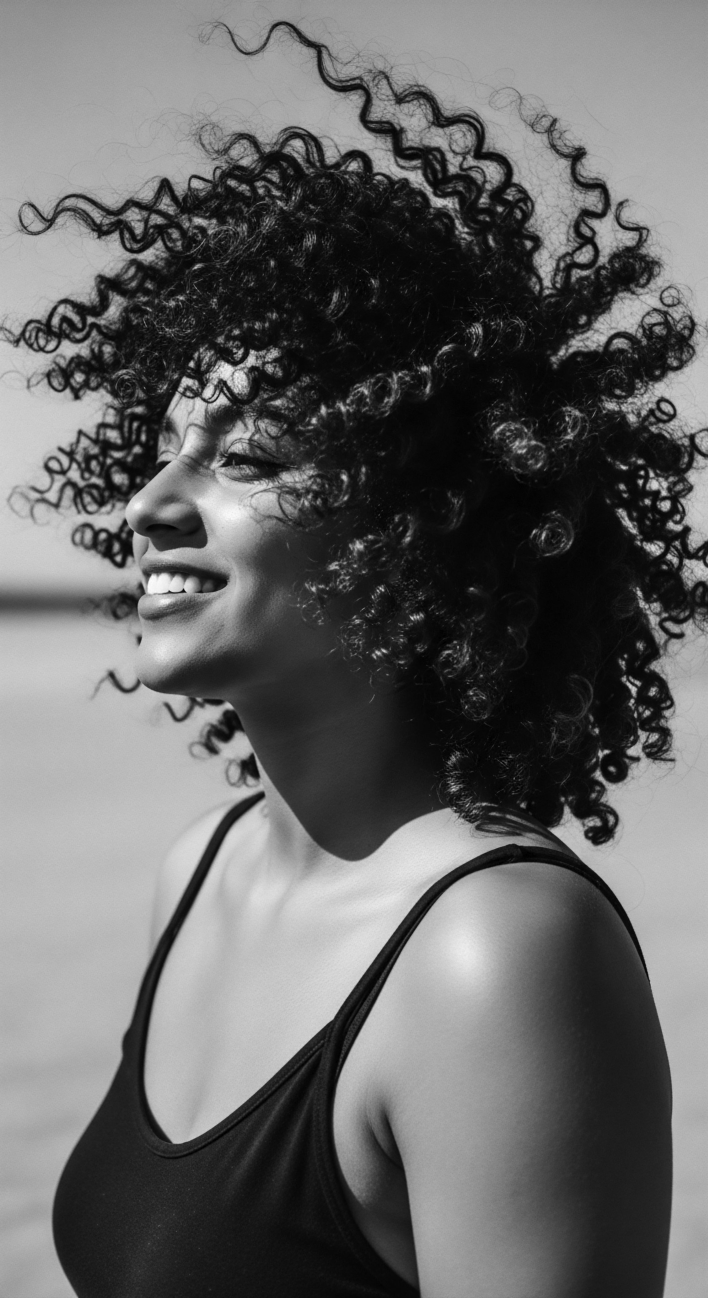 Woman with Voluminous Afro Textured Coils Smiling in Sunlight Black and White Bathed in sunlight, she exudes joy and confidence a testament to the beauty of afro texture. Her authentic smile paired with the wild freedom of her coils evokes a celebration of natural black hair heritage and embrace self love through ancestral genetic heritage and the freedom of expression.