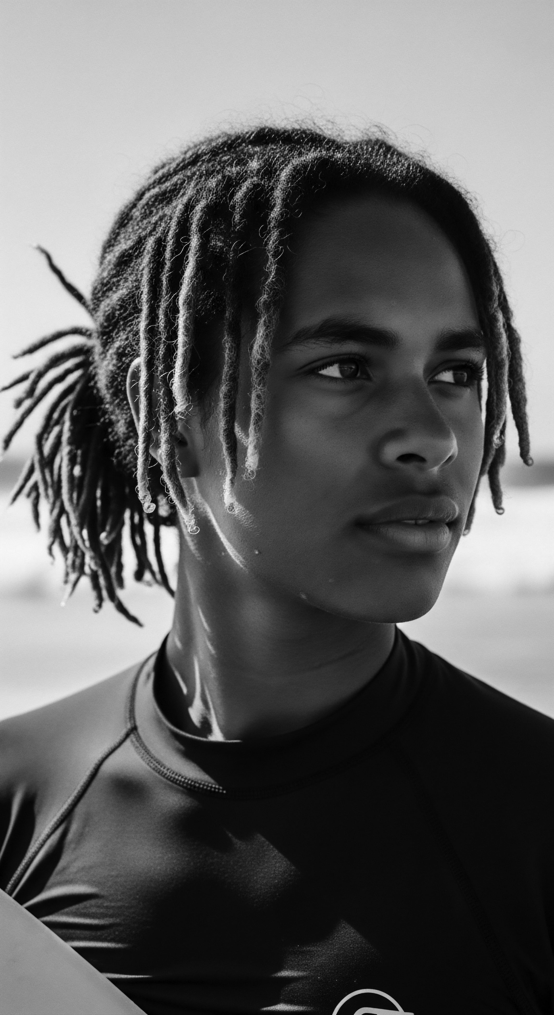 Captured in sharp monochrome, the serene gaze of a youth with styled locs evokes themes of identity and ancestral heritage while reflecting a commitment to holistic hair care and expressive styling. The surf backdrop subtly hints at harmonious connection with nature, enriching the cultural narrative.